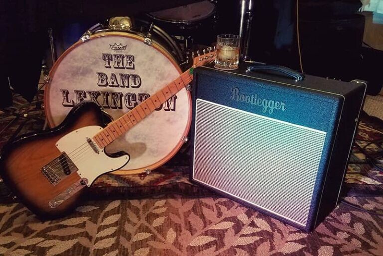 a honey color guitar and bootlegger guitar amp is kept on the floor with other musical instruments